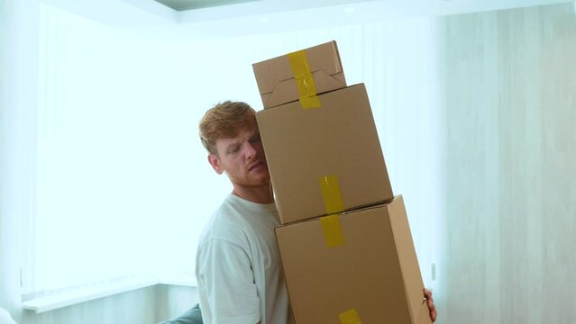 Close up of handsome Caucasian red-haired guy holding many cardboard boxes in hands. Boxes falling on the floor. Moving out the apartment. Relocation concept. Slow motion, indoors