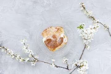 Fototapeta na wymiar Choux Bun with whipped cream and sugar powder on top on glass plate. Gray background with spring white cherry blossom flowers. Delicate choux pastry dessert. French cream puff. Top view, copy space