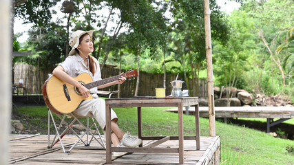 Hipster young woman playing acoustic guitar during camping near the river bank. Outdoor activity or vacation concept