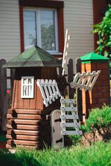 A small decorative wooden dark windmill with while blades and a small white window. Placed against a modern house. Grass and a bush on the foreground