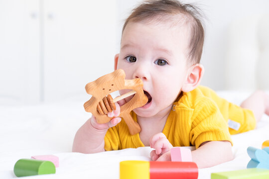 healthy asian baby girl in yellow bodysuit plays with wooden toy teether for teeth on white bedding