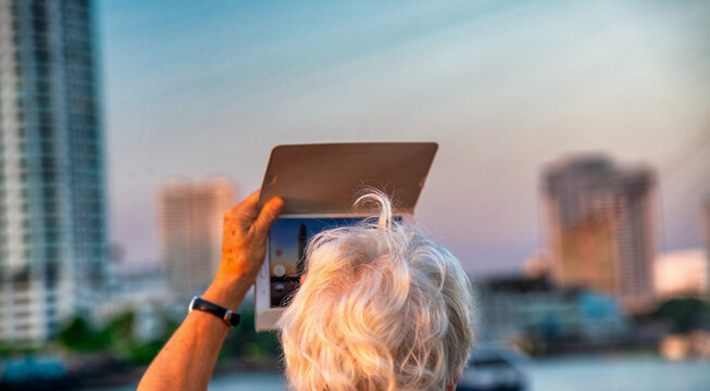 Elderly woman taking pictures of beautiful city skyline at sunset with tablet.