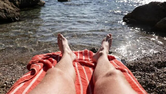 Man sunbathing by the sea between rocks on the beach. Leg shot of man sunbathing on red and white stripped towel with the sun beating off the water.
