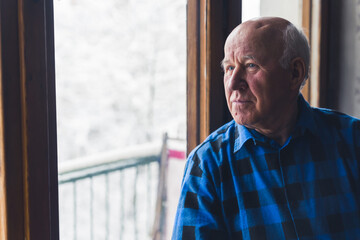Bald caucasian old senior man standing by the window on a winter day and looking away. High quality...