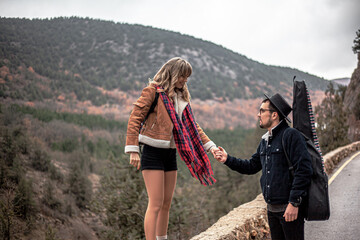 Portrait of couple in love kissing in the countryside road against the mountain and road, man holding the guitar, people hitchhiking