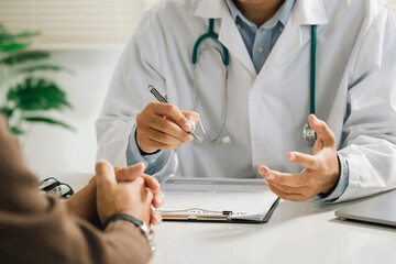 A professional physician in a white medical uniform talks to discuss results or symptoms and gives a recommendation to a male patient and signs a medical paper at an appointment visit in the clinic. - obrazy, fototapety, plakaty