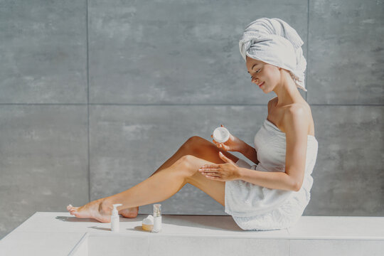 Horizontal shot of pretty woman with slender legs puts moisturising cream on healthy skin enjoys beauty routine after taking bath poses in bathroom wrapped in towel enjoys softness after lotion