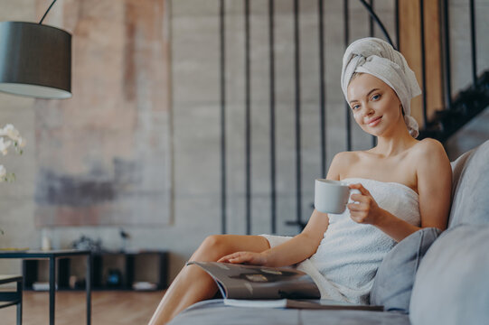 Pleased relaxed woman has rest at home poses on comfortable sofa wrapped in bath towel reads magazine drinks coffee poses over cosy home interior feels refreshed and pleased. People leisure.