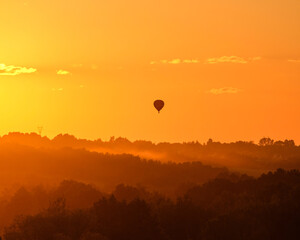 Silhouette of a hot air balloon flying over the treetops in an orange sky at sunset.