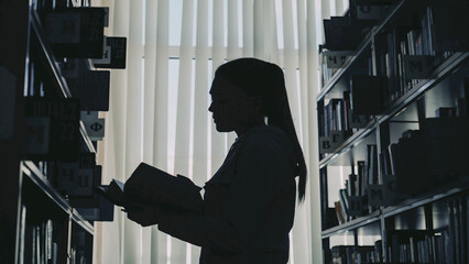 Silhouette of young student girl standing among bookshelves in big university library against...