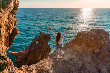 A female tourist, standing with her hands up, reaches the ends of the earth and sees the dragon rock. The girl welcomes the sun. Concept design. A successful woman looks at the sunset with open arms.