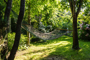 Wicker hammock, stretched and fixed on tree trunks. Blurred background of landscaped garden greenery. Close-up. Celebration atmosphere. Bright sunny day. Serene rest and relaxation.