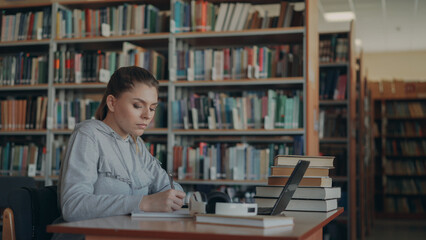 Beautiful female student sitting at table with books in library writing down summary in copybook...