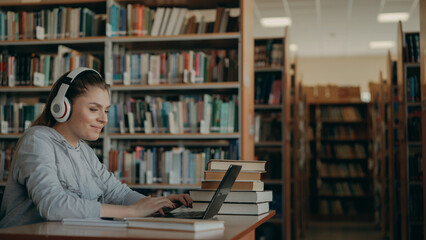 Beautiful positive caucasian female student with big white headphones on head working at table in spacious library in front of laptop. She is smiling and texting