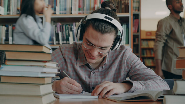Young handsome caucasian schoolboy sitting at table in big college library is writing something in copybook. He is smiling and looks positive and happy, books lying on table