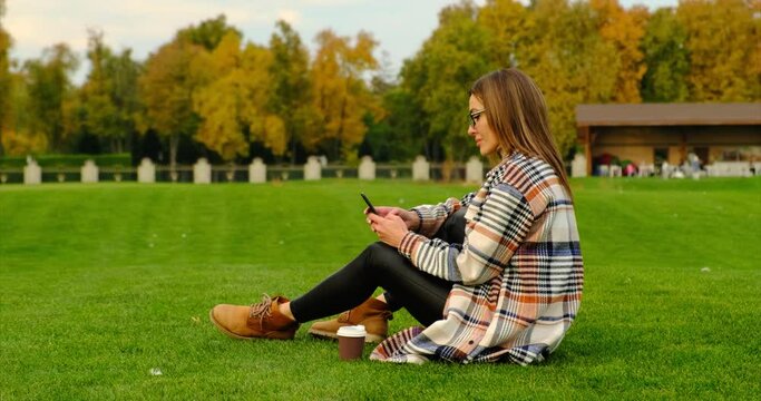 beautiful woman holding coffee sitting on the Grass. stylish english girl in coat sits on green grass in central park and enjoys hot coffee. girl enjoying takeaway coffee cup on sunny cols fall day