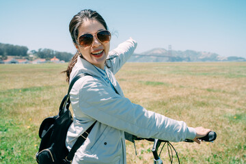 portrait happy asian girl on bike is pointing into the distance while looking at the camera with a smile on a green lawn under sunny blue sky.