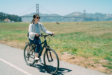 asian woman wearing sunglasses is having summer holiday fun by riding along a green field at leisure with golden gate bridge at background in California USA.