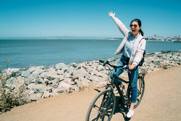 cheerful asian woman is raising arm and shouting with joy into distance on bike while visiting the...