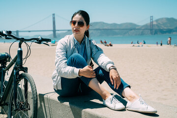 trendy chinese female is looking into the distance while sitting by her bicycle taking a break on the stone pier at the beach in San Francisco USA at summertime