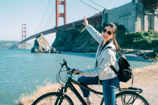 Happy Asian Girl Tourist Is Looking At The Camera With A Peace Sign Hand Gesture On Bike With Golden Gate Bridge And Blue Sea At Background In San Francisco USA