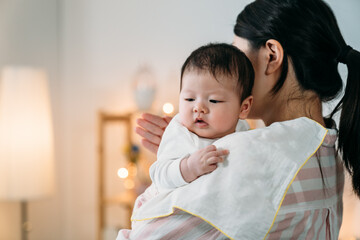 closeup innocent asian infant baby leaning on her mom’s shoulder with cloth is looking away into the space while her mom is burping patting on her back at home. - obrazy, fototapety, plakaty