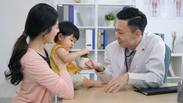 friendly pediatrician is playing with the cute baby patient by touching her with finger and giving her a pot plant during a consultation in the office