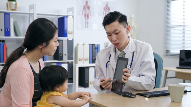 asian pediatrician is tapping the screen with a pen while explaining the test results to the mother and her kid on a touchpad in his clinic office.