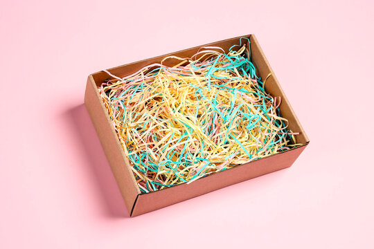 Open brown box with colorful pastel shredded paper on pink background.