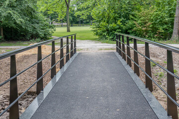 New black metal bridge placed over a stream, footpath leading to a public park with green grass, lush trees in the background, sunny spring day in Heerlen, South Limburg, Netherlands