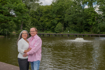 Romantic scene of mature couple standing hugging, looking at camera, pink and white casual clothes, pond with a fountain, trees in background in public park in Heerlen, South Limburg, Netherlands