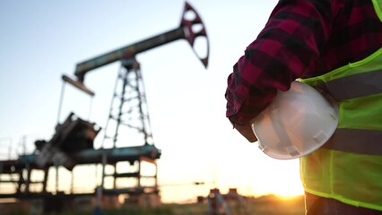 oil production. a worker holding a protective hard hat at sunset in the background an oil pump....