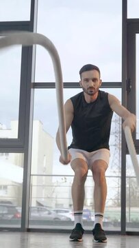 vertical video of concentrated sportsman doing exercise with battle ropes in gym