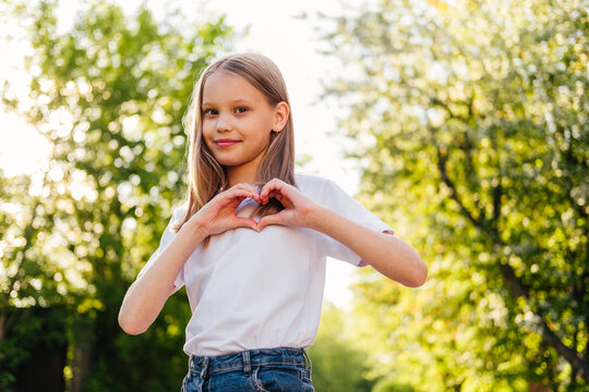 Smiling girl showing heart sign with hands outdoor. Child making heart gesture at sunset. Health and love concept