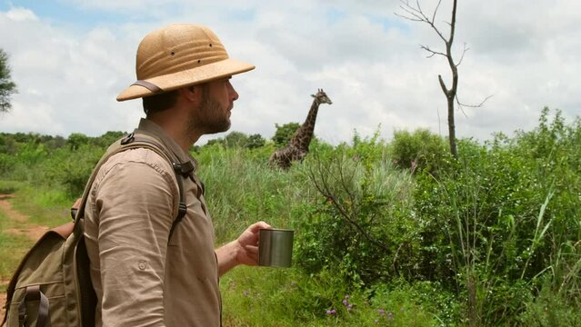 man traveler with backpack drinking water while standing on mountain. A male drink water in sand desert safari. a man zoologist drinks water from a tourist iron mug and watches a giraffe