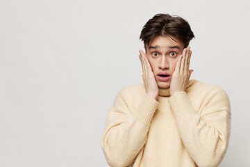 Portrait photo. Horizontal studio shot. close-up photo.An emotional young man with dark, short, beautiful hair combed back in a beige sweater with a high neck stands on a gray background with his
