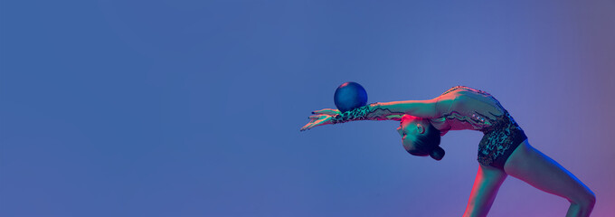 Portrait of young flexible girl, rhythmic gymnast athlete training with blue ball isolated on blue purple background in neon light