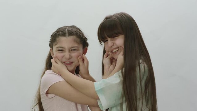 2 little girlfriends who love each other very much pinch each other's nose and cheeks on a white background. Portrait of friends who love each other very much.