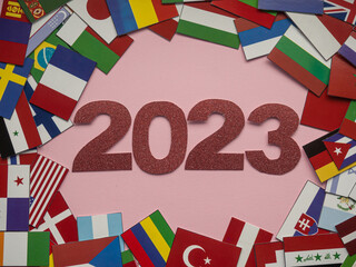 2023 on the background of many flags. 2023 red figures lie next to small flags of different countries on pink backdrop. Christmas and New year card. Multinational concept of celebrating winter holiday