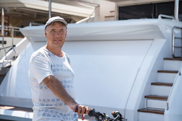Senior man posing against the background of a white yacht while riding a bike on the beach