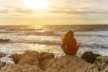 The girl sits on a stone and looks at the sea. Sunset on the sea, a lonely woman enjoys the sunset.