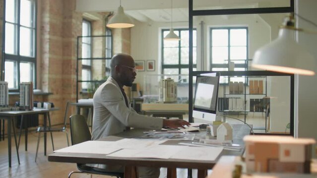 African American male architect typing on computer at desk, then standing up and walking away during workday in office