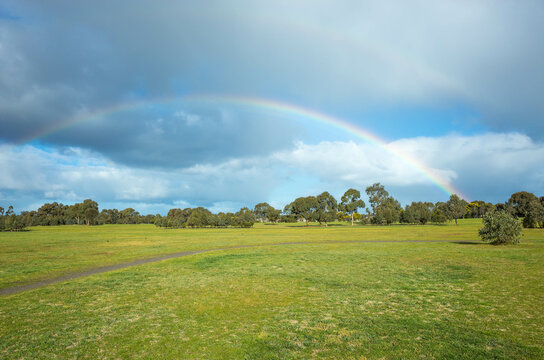 A large park with double rainbows against cloudy sky. Background texture of green grass field or meadow with Australian native trees and a small footpath. Copy space for text. Presidents Park Werribee