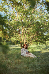 Pretty red haired young woman in summer dress is sitting under the tree and looking at phone in the rays of sun.