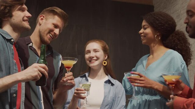 Medium slowmo of five young cheerful diverse LGBTQ friends chatting and drinking beer and cocktails while having outdoor terrace party on summer day