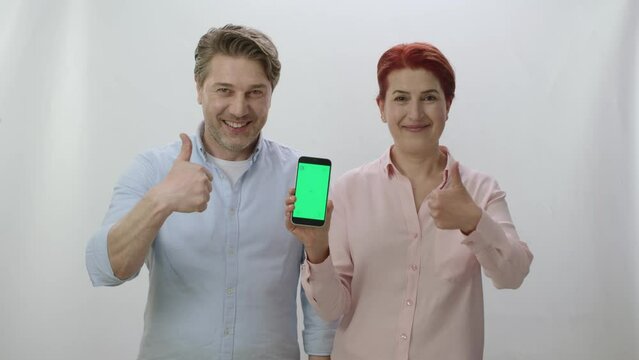 A couple is holding a smartphone with a green screen on a white background. Couple pointing at green screen smartphone giving thumbs up sign. You can place the image you want on the green screen.