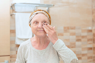 Mature woman standing at the mirror in the bathroom and putting a scrub on her face. Elderly woman taking care of her face