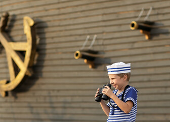 A boy of 7-8 years old dressed as a sailor sits in a wooden brown barrel. The boy is playing with marine binoculars. Child vacations in the park. The model of a sailing ship is standing.