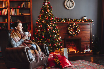 Girl at Home by Christmas tree in Warm Scarf Looks out Window. Woman Holding Cup of Aromatic Drink