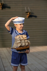 A boy of 7-8 years old dressed as a sailor sits in a wooden brown barrel. The boy is playing with marine binoculars. Child vacations in the park. The model of a sailing ship is standing.